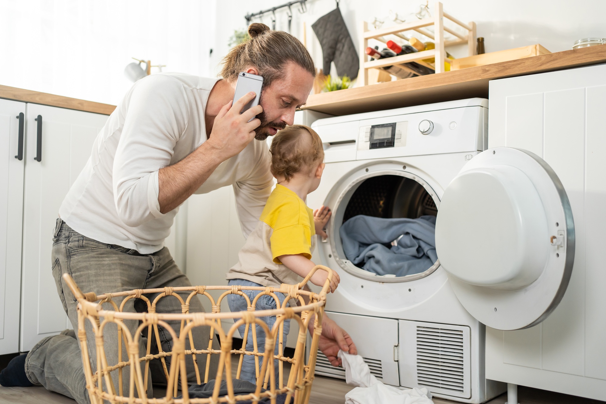 Caucasian busy father doing housework with baby boy toddler in kitchen.