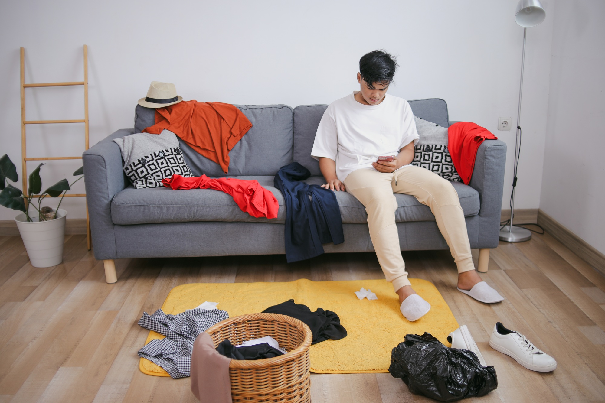 Young Man Sitting On Sofa Playing Smartphone With Lots Of Clothes Mess The Room
